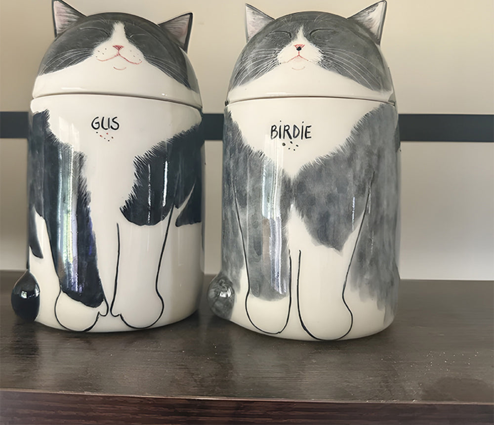 Two ceramic cat-shaped jars with 'GLIS' and 'BIRDIE' on a wooden surface.
