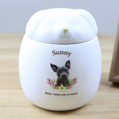White pet urn with a dog's face and 'Sunny' printed on it, placed on a wooden surface.