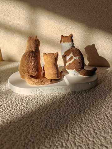 Three cat figurines on a white base against a beige wall.