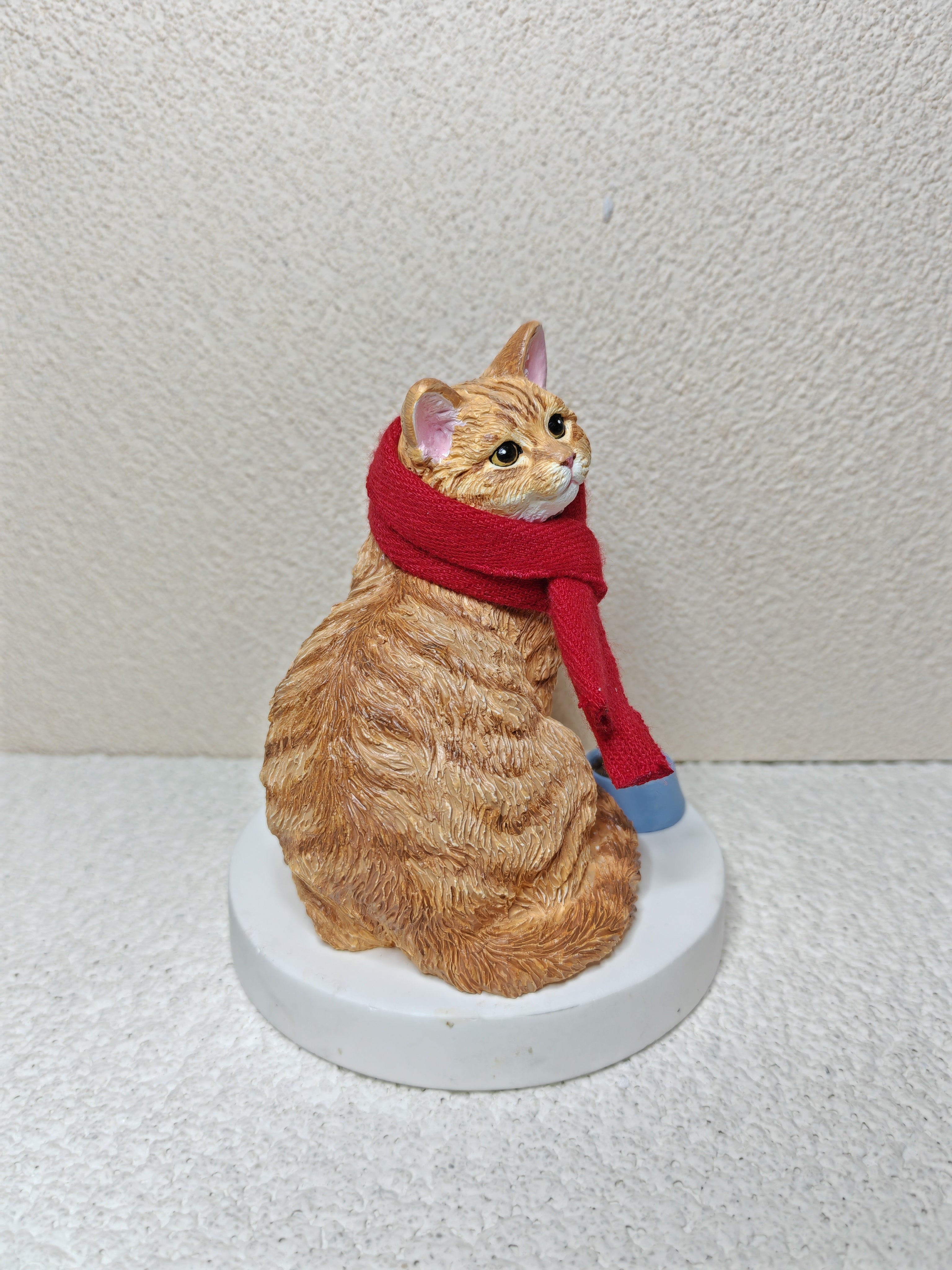 Cat figurine wearing a red scarf on a white pedestal against a plain background