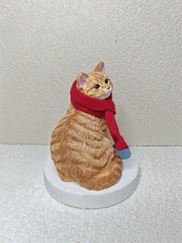 Cat figurine wearing a red scarf on a white pedestal against a plain background