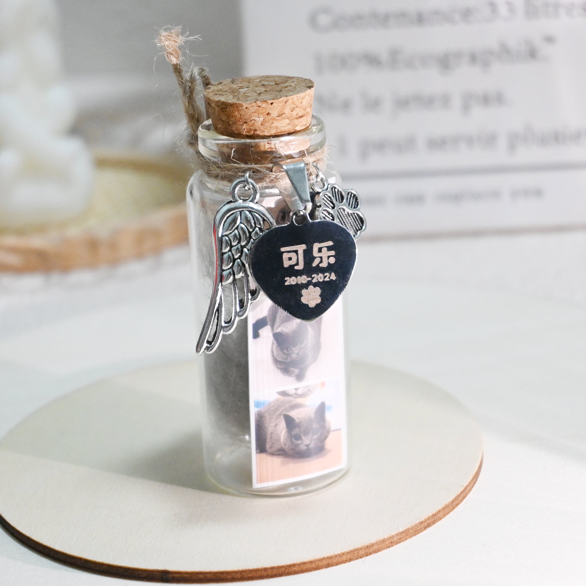 Glass bottle with cork lid containing a photo of a cat, angel wing charm, and heart-shaped tag on a white surface.