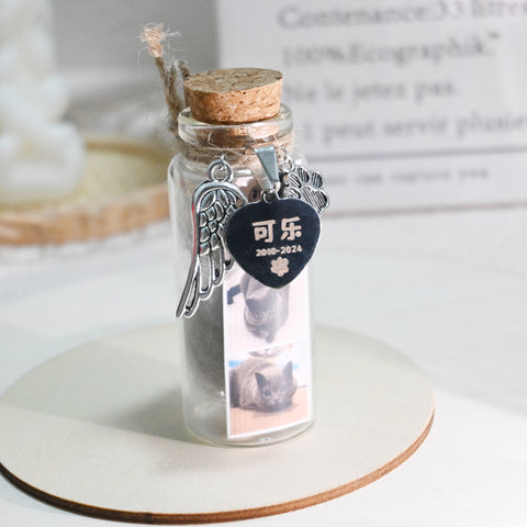 Glass bottle with cork lid containing a photo of a cat, angel wing charm, and heart-shaped tag on a white surface.