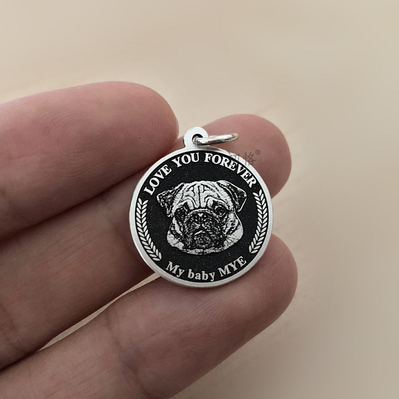 Silver pendant with pug illustration and text held between fingers on a beige background