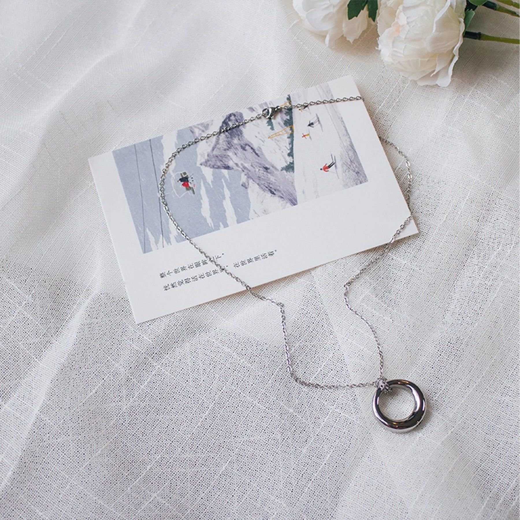 Silver necklace with a circular pendant on a white card with illustrations, placed on a textured white surface.