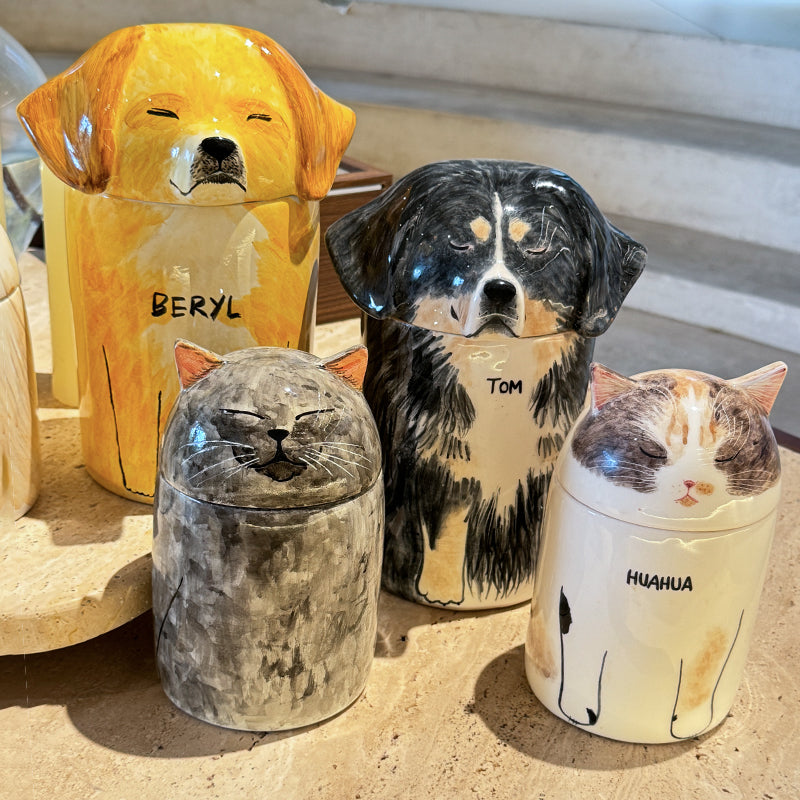 Four ceramic containers shaped like dogs and cats with names on a sandy surface.