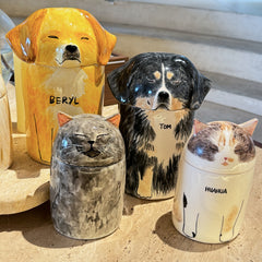 Four ceramic containers shaped like dogs and cats with names on a sandy surface.